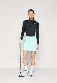 Černý golfový top s dlouhým rukávem a čtvrtzipem, zkombinovaný s lehce mátově zelenou sukní. Bílé golfové boty a ponožky, drží golfovou hůl.