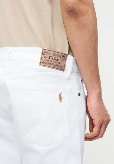 Κοντινό πλάνο ενός ατόμου που φοράει λευκά τζιν Polo Ralph Lauren με καφέ λογότυπο και κεντητό έμβλημα αλόγου στην πίσω τσέπη.