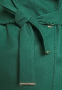 Groene, gestructureerde stof met een gestrikte riem. Bevat een goudkleurige accent en heeft een glad oppervlak met nette stiksels langs de randen.