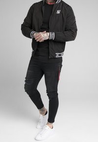 Muž v černé bomber jacket, černých roztrhaných skinny džínech, bílých teniskách, který ukazuje tetované ruce a krk na jednobarevném pozadí.