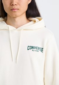 Sudadera con capucha de color crema hecha de una tela suave. Presenta el texto verde "CONVERSE ALL*STAR" en el pecho. Cuerdas ajustables en el cuello.