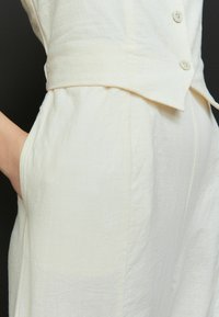 Robe en lin blanc avec un corsage ajusté, un col en V et trois boutons. Dispose de poches latérales et d'un tissu légèrement texturé.