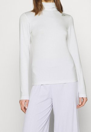 Femme portant un haut blanc ajusté à manches longues et col roulé avec un petit logo sur la partie supérieure gauche de la poitrine et un pantalon clair et ample.