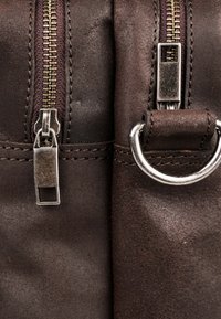 Detalle de un bolso de cuero marrón con dos cremalleras metálicas, herrajes plateados y un acento de anillo circular, destacando la textura y las costuras.