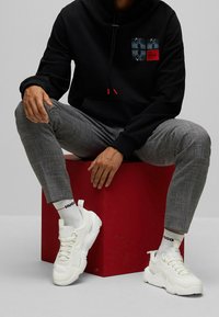 Černá mikina s kapucí a přední kapsou, šedé kostkované kalhoty a robustní bílé tenisky. Červené akcenty na mikině a ponožkách s logem. Sedící na červené krabici.