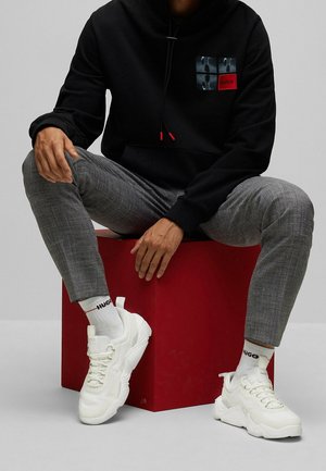 Haut à capuche noir avec une poche avant, pantalon à carreaux gris et baskets blanches épaisses. Accents rouges sur le haut à capuche et les chaussettes avec logo. Assis sur une boîte rouge.