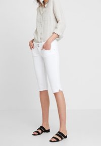 Pantalones blancos hasta la rodilla con aberturas laterales, combinados con una camisa de lino beige clara. La camisa tiene una tapeta con botones y mangas largas.