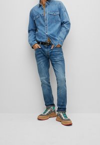 Chaqueta de mezclilla y jeans en azul claro, combinados con deportivas gruesas en beige y gris con acentos turquesas y suelas rojas. Cinturón negro incluido.