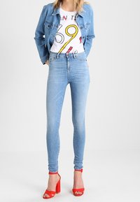 Ljust blå höga jeans och en vit grafisk t-shirt med röda och gula accenter, tillsammans med röda klacksandaler. Denimjacka bärs.