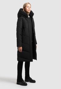 Abrigo acolchado largo y negro con un cuello alto, que presenta un patrón de quilt diagonal y botones en el frente. Llevado sobre pantalones negros y botas.