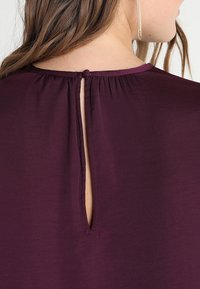 Vue arrière d'une femme portant un blouse couleur prune avec une fermeture à clé boutonnée au niveau du cou et de longs cheveux bruns.
