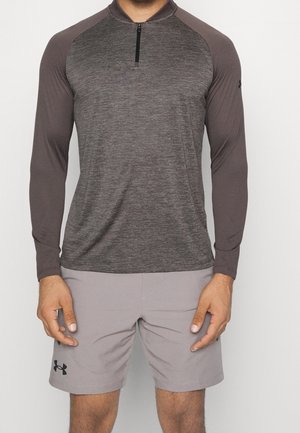 Mand iført en langærmet, mørkegrå trøje med lynlås og lysegrå træningsshorts, står med armene afslappet ned langs siderne.