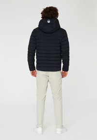Marineblauwe pufferjack met horizontaal gestikt ontwerp en een verstelbare capuchon, gecombineerd met lichte broeken en witte sneakers.