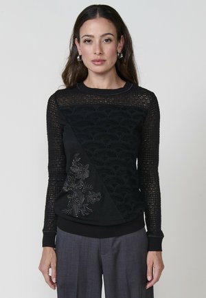 Une femme se tient debout, portant un pull noir avec des motifs intriqués et des détails floraux en perles, associé à un pantalon gris. Elle a de longs cheveux ondulés.
