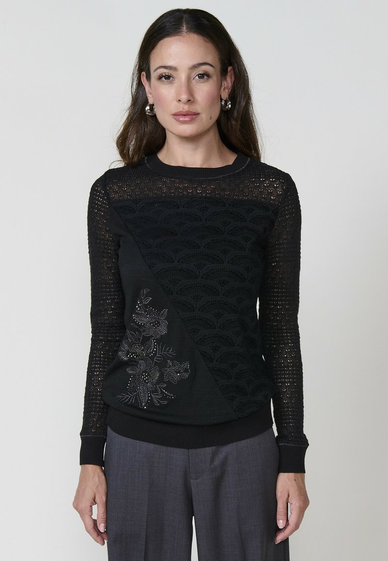 Una donna è in piedi indossando un maglione nero con intricati motivi e dettagli floreali in perline, abbinato a pantaloni grigi. Ha i capelli lunghi e mossi.
