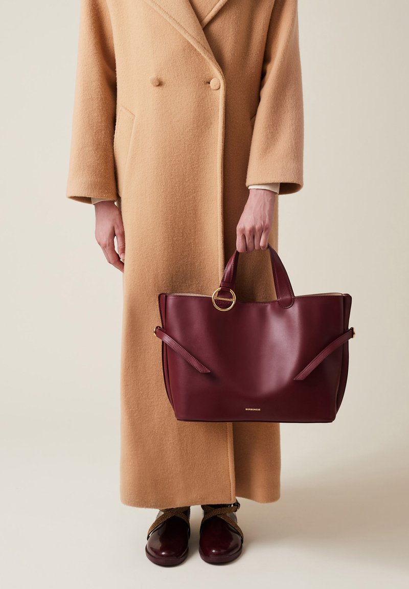 Burgundy leather tote bag with dual handles, gold ring accent, and side straps, held by a person wearing a beige overcoat and brown shoes.