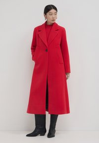 Femme portant un long manteau rouge à boutons, un pull rouge, et des bottes noires à genoux embossées, se tenant devant un fond blanc.
