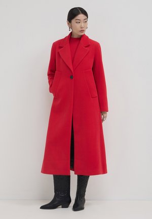 Woman wearing a long red buttoned coat, red sweater, black embossed knee-high boots, standing against a white background.
