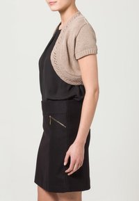 Beige stickad bolero med korta ärmar och en vågig fåll. Matchad med en svart kjol med en dragkedjeficka framtill.