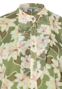 Blouse verte avec un motif floral en rose et beige. Elle présente une patte à volants et une fermeture à boutons avec des boutons ronds assortis.