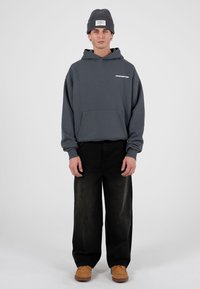 Felpa grigia con cappuccio e tasca frontale, con testo bianco. Abbinata a jeans neri larghi e scarpe colorocuoio. Il modello indossa un berretto di maglia grigio.