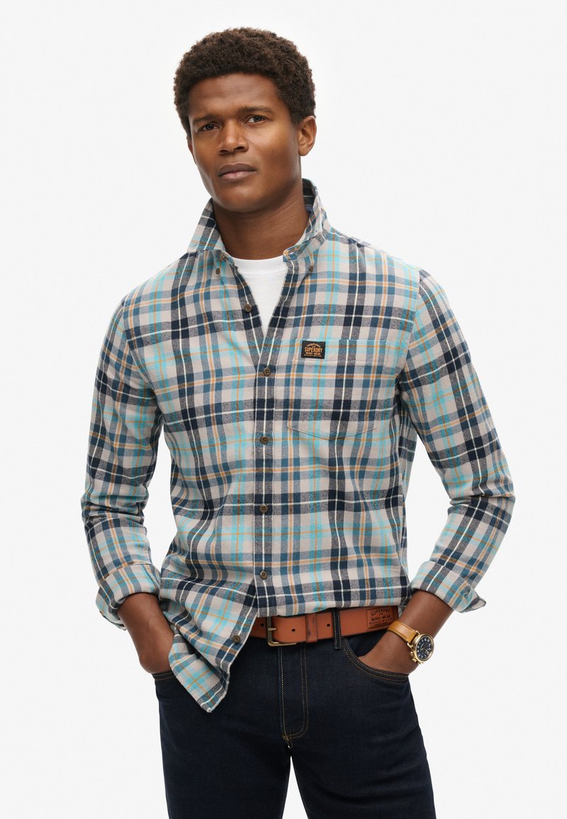 Camisa a cuadros en colores azul, beige y marino, con collar abotonado, bolsillo en el pecho y parche de la marca. Llevada con un cinturón marrón.