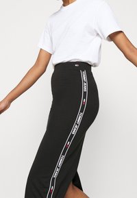 Černá midi sukně s bočním rozparkem, s bílým potiskem "TOMMY JEANS" podél levé strany a přiléhavým pasem pro štíhlý siluetu.