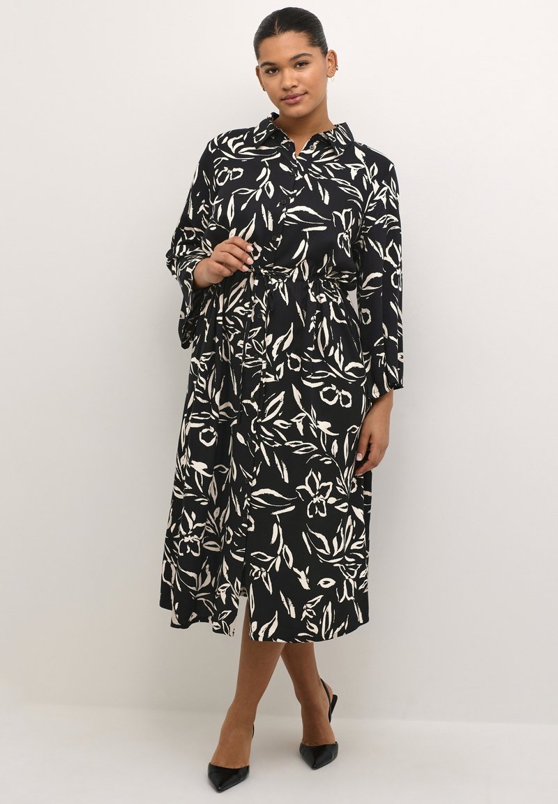 Schwarzes Kleid mit weißem Blumenblattmuster, Knopfleiste vorne, Kragen, langen Ärmeln, elastischem Bund und knöchellangem Saum. Kombiniert mit schwarzen High Heels.