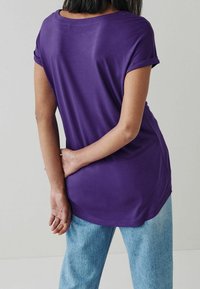 Lila T-Shirt mit umgeschlagenen Ärmeln, lockerer Passform und abgerundetem Saum; kombiniert mit hellen Jeans. Glatte Stofftextur.