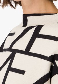 Gebreide trui met een hoge hals, crèmekleurige stof met een gedurfd zwart geometrisch patroon en een zachte textuur. Close-up van het schoudergebied.