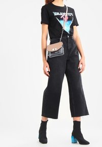 T-shirt graphique noire avec un design multicolore, jean noir taille haute à jambes larges, sac transparent avec accents beiges, et bottines noires à talons bleus.