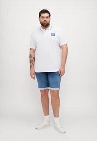 Camisa polo blanca con detalles y logo en color azul, llevada con shorts de mezclilla azul claro, combinada con zapatillas blancas y calcetines hasta los tobillos.