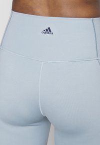 Jasnoniebieskie legginsy z wysokim stanem o gładkiej teksturze, z granatowym logo Adidas i spłaszczonymi szwami dla zwiększenia komfortu.