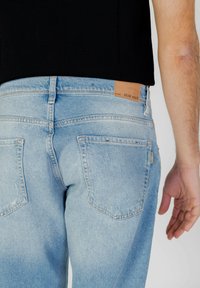 Jeans de mezclilla azul claro con corte recto, que presentan una etiqueta de cuero rectangular en la cintura y los bolsillos traseros, además de ligeros detalles de desgaste.