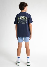Marinblå bomullströja med en stor grön och vit grafik som står "PETROL" på ryggen, tillsammans med ljusblå shorts och vita sneakers.