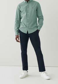 Zielono-biała w paski koszula z guzikami, granatowe spodnie i białe sneakersy. Koszula ma małe haftowane logo na piersi.