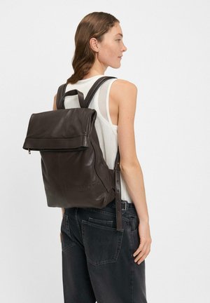 Frau in einem ärmellosen weißen Oberteil und schwarzen Jeans, die einen großen dunkelbraunen Leder-Rucksack mit umklappbarem Deckel trägt.