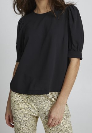 Kobieta ubrana w czarną bluzkę z bufkami, w połączeniu z jasnobeżowymi spodniami z wysokim stanem, ozdobionymi małym czarnym wzorem w kropki.