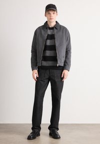 Young man wearing a black cap, grey zip-up jacket, black and grey striped sweater, black pants, and black shoes standing against a white wall.