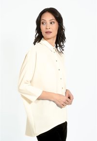 Kremkrāsas blūze ar pogu aizdari priekšpusē, apkakli un trīs ceturtdaļu piedurknēm. Izgatavota no gludas, vieglas auduma ar brīvu piegriezumu.