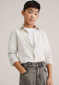 Világos bézs, gombos ing klasszikus gallérral, feltekeredett ujjakkal, fehér póló felett viselve. Magas derekú szürke farmernadrággal párosítva.