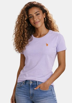 Junge Frau mit langen lockigen Haaren, trägt goldene Creolen, ein hellviolettes T-Shirt mit kleinem orangem Logo und blaue Jeans, eine Hand in der Hosentasche.