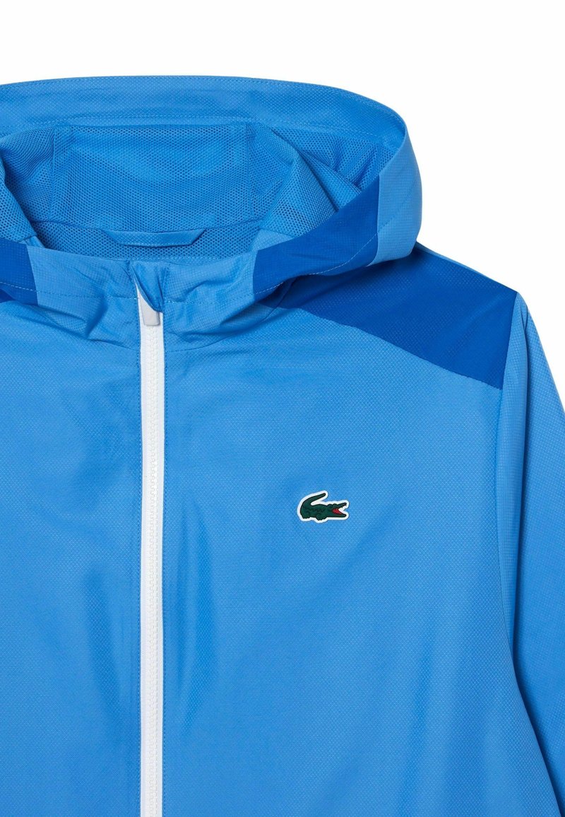 Coupe-vent bleu clair avec une capuche, doté d'une fermeture éclair blanche et d'accents d'épaule bleu foncé. Logo Lacoste affiché sur la poitrine.