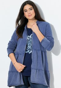 Felpa blu con zip e orlo arricciato, realizzata in tessuto morbido. Presenta maniche lunghe e cappuccio. Indossata sopra una maglietta blu scuro con un motivo floreale.