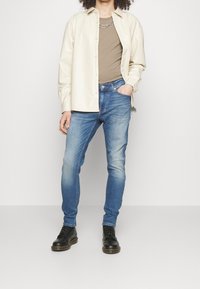 Béžová košile, taupe tričko, modré džínsy, černé šněrovací boty. Kcasualní outfit s volným střihem a zapínáním na knoflíky.