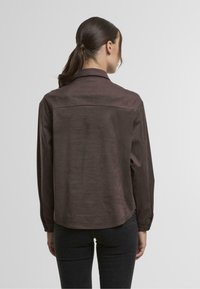 Brown, button-up shirt with a smooth texture, long sleeves, and a rounded hem, featuring a back yoke and subtle sheen.