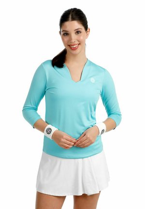 Jeune femme en haut de sport bleu clair à manches longues et jupe blanche, portant des poignets blancs, souriant les mains jointes devant elle.