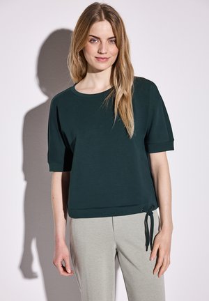 Dark green short-sleeve top with a round neckline and a tied accent at the hem. Smooth fabric, relaxed fit, paired with light gray pants.