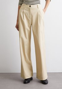  hooggehaalde, wijde beige broek met voorplooien, zijzakken en een knoopsluiting. Draag met een zwart-wit gestreept topje en zwarte schoenen.