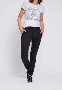 Svarta träningsbyxor med sidofickor och elastisk midja, tillsammans med en vit t-shirt med en svart abstrakt logotyp. Skorna är svarta sneakers.